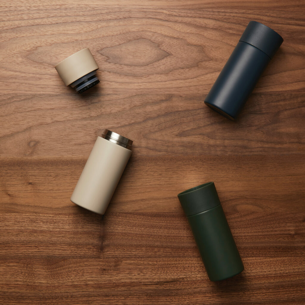 tre tazze termiche sdraiate su piano di legno: una color panna aperta, una verde e una grigia antracite